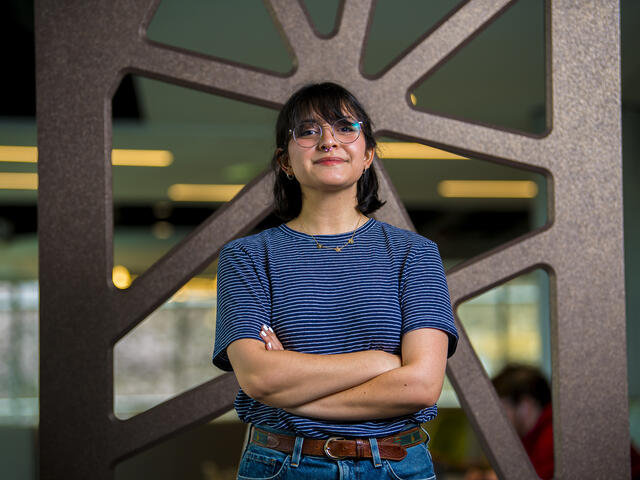 A student stands in front of a metal background with a blue shirt and arms crossed.