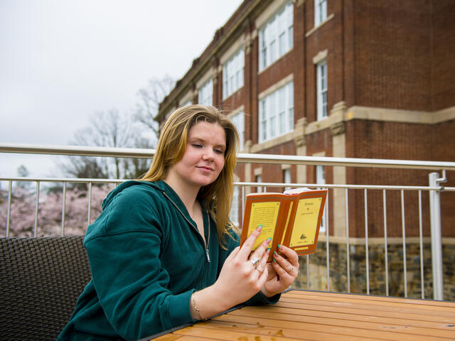 A student sits outside at a table while reading a book.