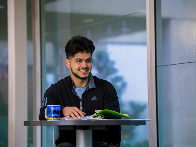 A student sits at a table with a notebook and mug in front of him.