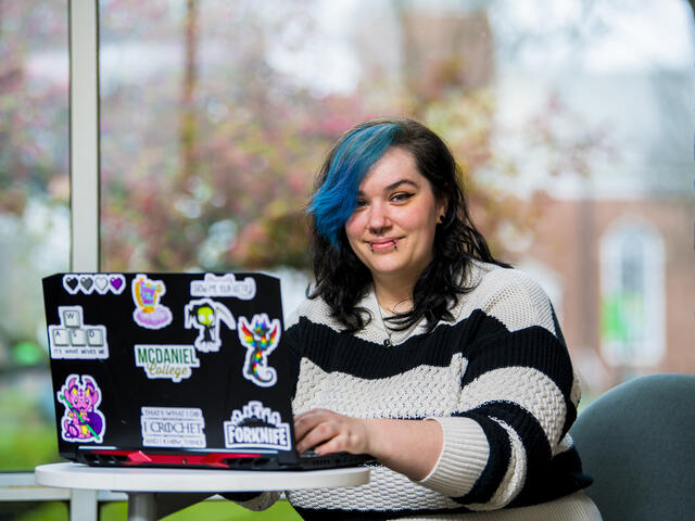 A student in a striped sweater sits in front of a window with a a laptop covered in stickers.