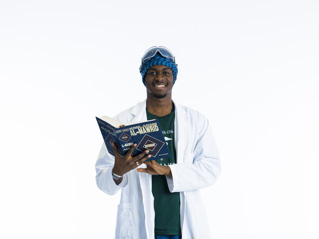 A student in a lab coat hold an Arabic book open while smiling at camera in front of a white background.