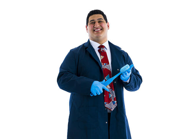A student in dark blue lab coat poses with giant toothbrush against white background.