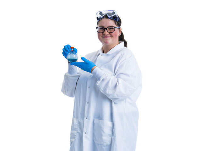 A student in a lab coat stands in front of a white background while wearing a pair of science goggles on their head and blue gloves and holding up a beaker with a blue liquid inside.