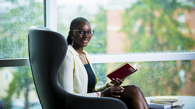 A students sits in a chair in front of a window while holding a book.