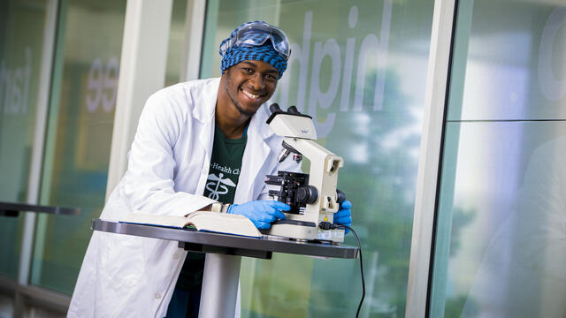 A student wearing a lab coat looks at a microscope while smiling.
