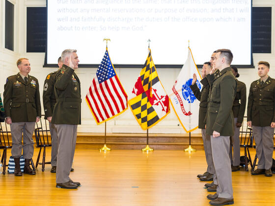 Photo of a stage with the U.S., Maryland, and Army flags and a row of cadets saluting an officer.