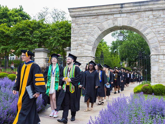 Students in regalia walk through the historical arch, led by the provost in regalia