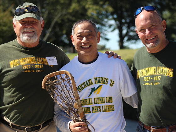 Three men stand together wearing memorial lacross t-shirts. The one in the middle holds an old lacrosse stick.