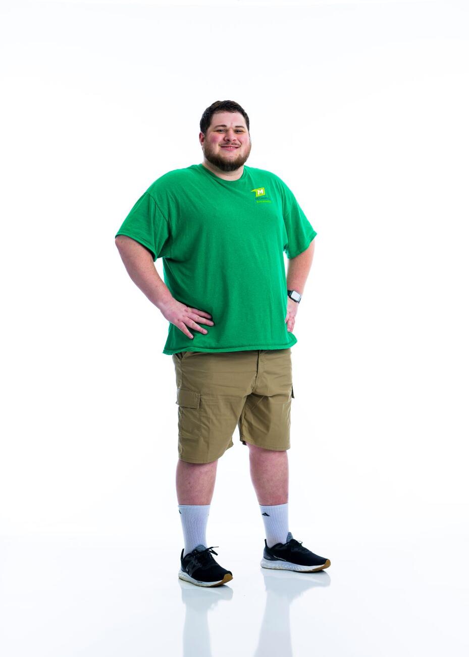 A student in a green shirt stands with his hands on his hips in front of a white background.