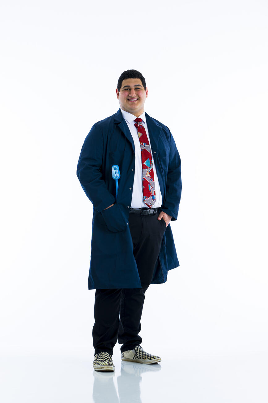 A student in dark blue lab coat with giant toothbrush in the pocket smiles at camera against white background.
