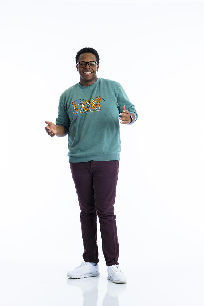 A student in a green sweatshirt with his arms out stands in front of a white background. 