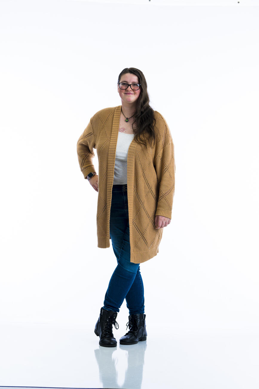 A student in a brown cardigan stands with one hand on her hip in front of a white background.