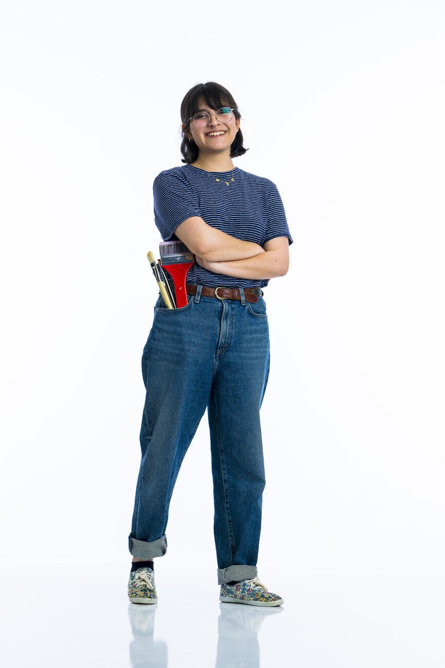 A student stands with her arms crossed and a set of paintbrushes in her pocket in front of a white background.