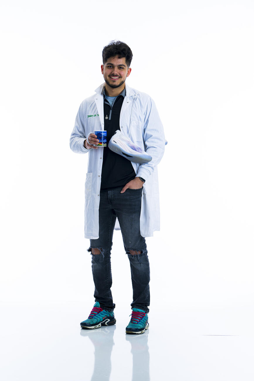 A student in a white lab coat stands holding a mug with basketball shoes over his shoulder in front of a white background.