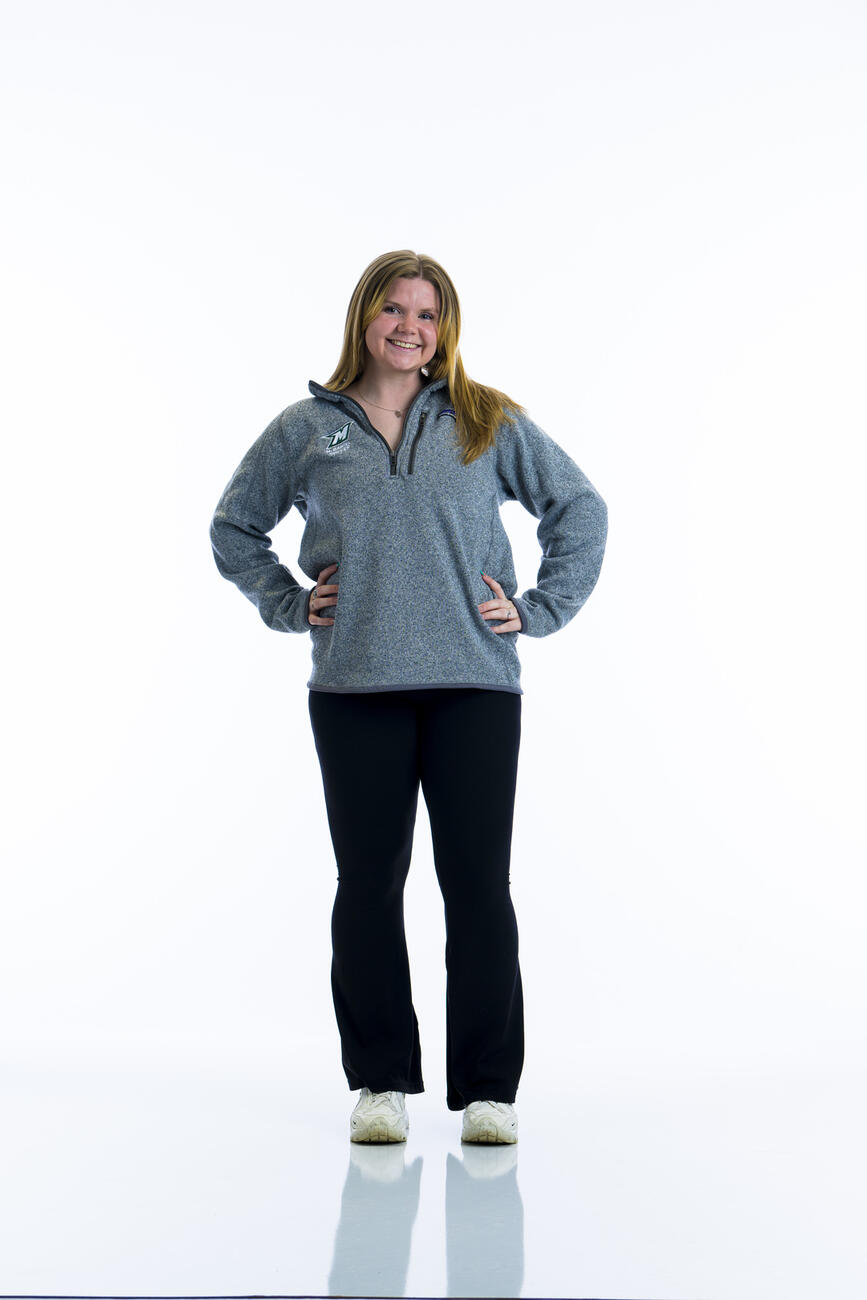 A student in a grey sweatshirt stands with her hands on her hips in front of a white background.