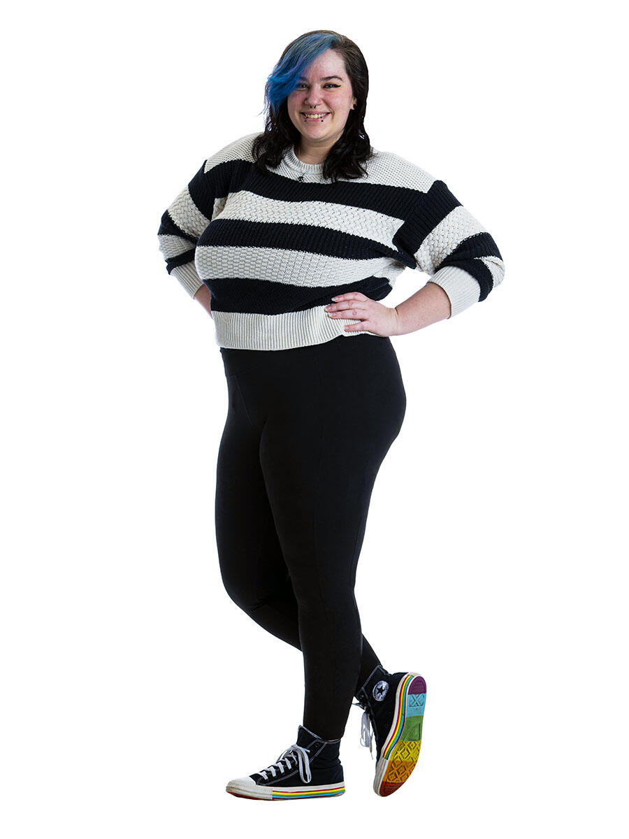 A student stands in front of a white background, wearing a black and white striped shirt and leaning on one leg to show a rainbow on the sole of their shoe.
