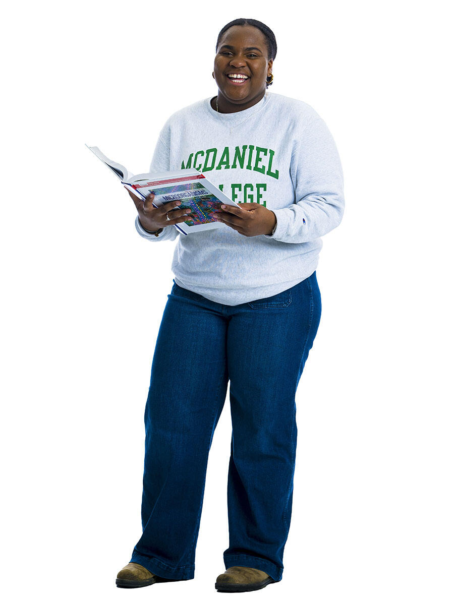 A student in a McDaniel sweatshirt stands in front of a white background while holding a textbook open in her hands.