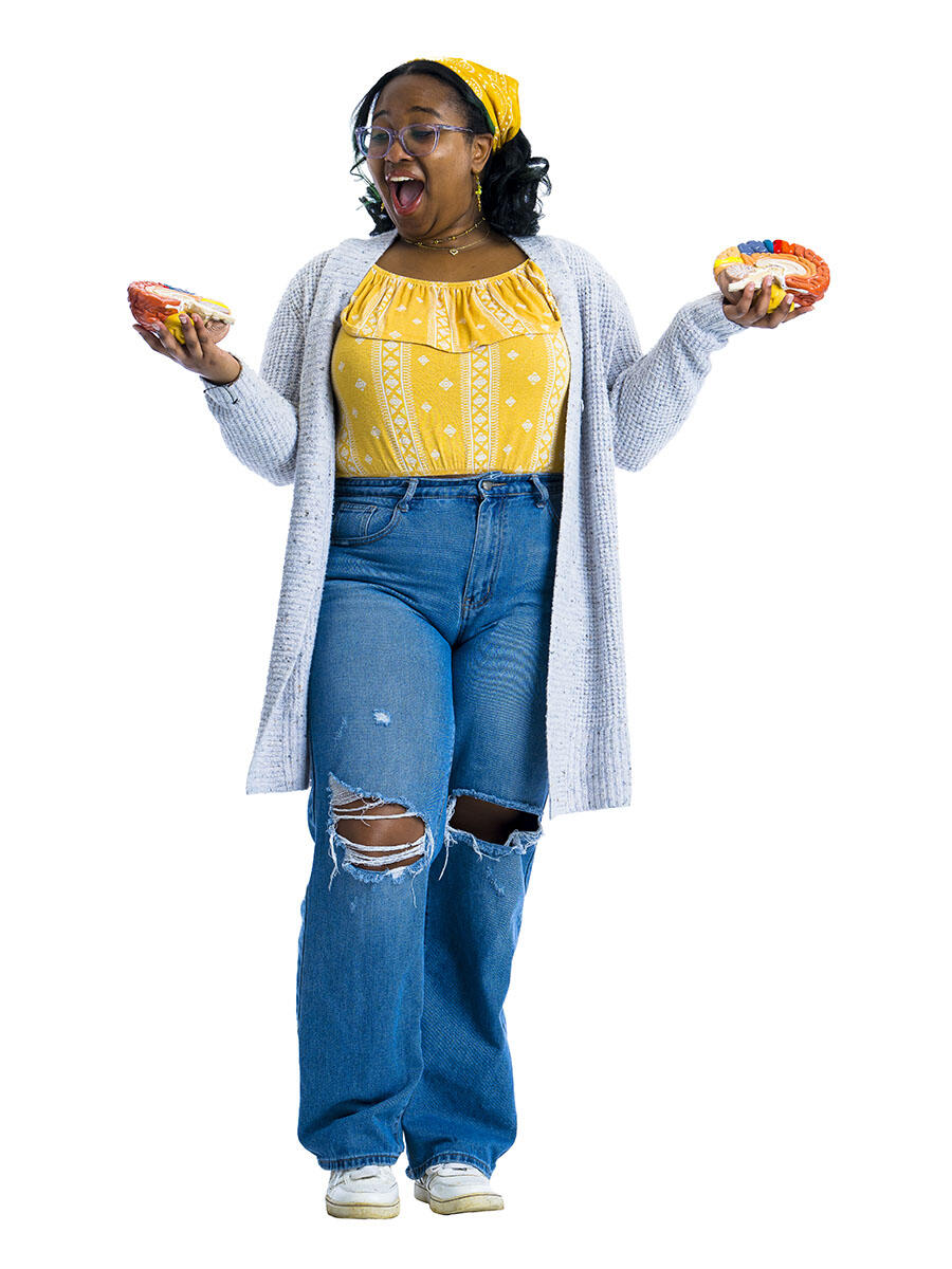 A student in a yellow shirt and jeans holds her hands out to either side while holding two halves of a model brain.