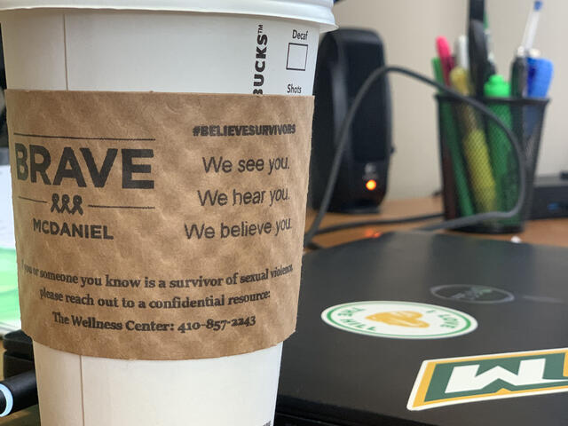 Starbucks coffee cup with BRAVE-McDaniel on paper coffee sleeve sitting on desk next to laptop.