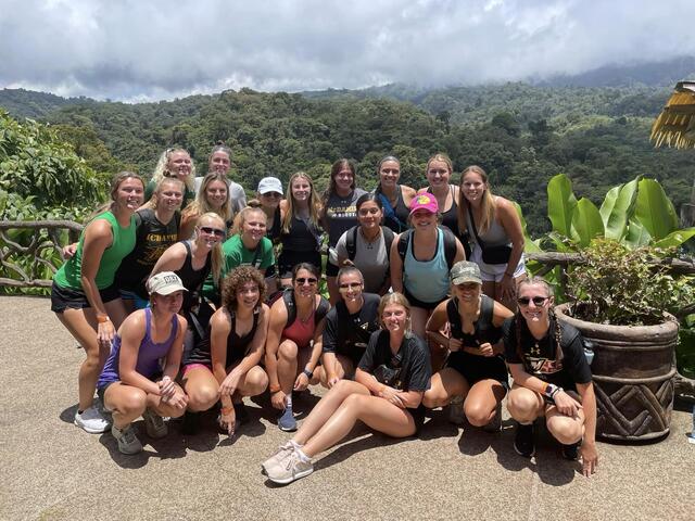 A group photo of the women's soccer team outdoors, in front of a tropical forest.