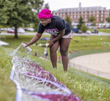 A female student leans over to spray paint on grass by the McDaniel football field with campus building in the background.