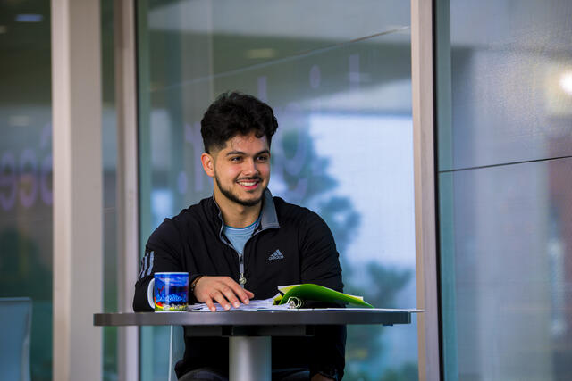 A student sits at a table with a notebook and mug in front of him.
