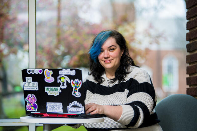 A student in a striped sweater sits in front of a window with a a laptop covered in stickers.