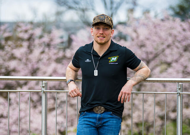 A student leans back on a railing in front of cherry blossom trees while wearing a camo hat and McDaniel wrestling shirt.