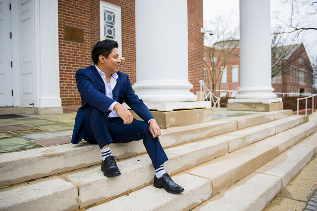 A student in a suit sits outside on steps in front of white columns.