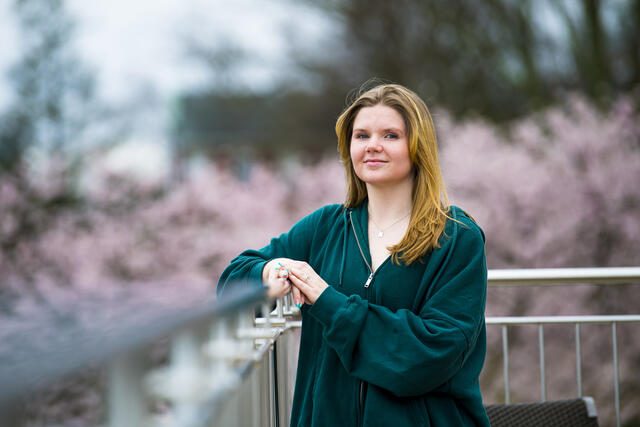 A student leans on elbow on an outdoor railing while wearing a green sweatshirt with pink cherry blossom tree behind them.