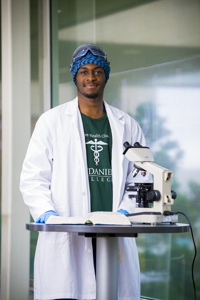 A student in a lab coat stands in front of a microscope and smiles at the camera.