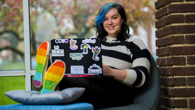 A student sits with a laptop on their lap and their feet up in front of them. The soles of their shoes are rainbow-striped.