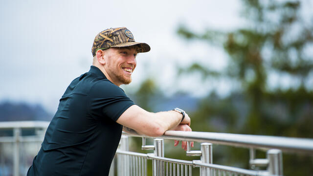 A student wearing a hat leans on a railing looking off camera while smiling.