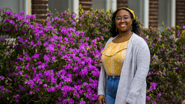 A student stands in front of a purple flowering bush outside while wearing a yellow shirt and yellow bandana.