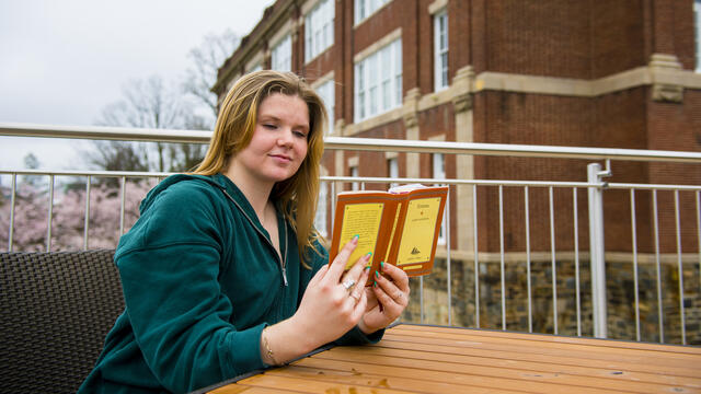 A student sits outside at a table while reading a book.