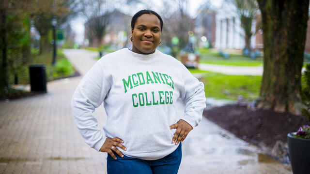 A student wearing a McDaniel College sweatshirt stands outside with her hands on her hips.