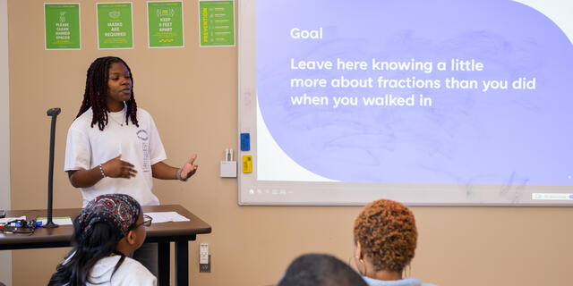 Photo of a Black student in front of a projector screen speaking to a classroom of people.
