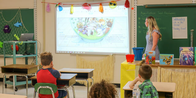 A female teacher looks at a projector image while standing at the front of a classroom with young children at desks.