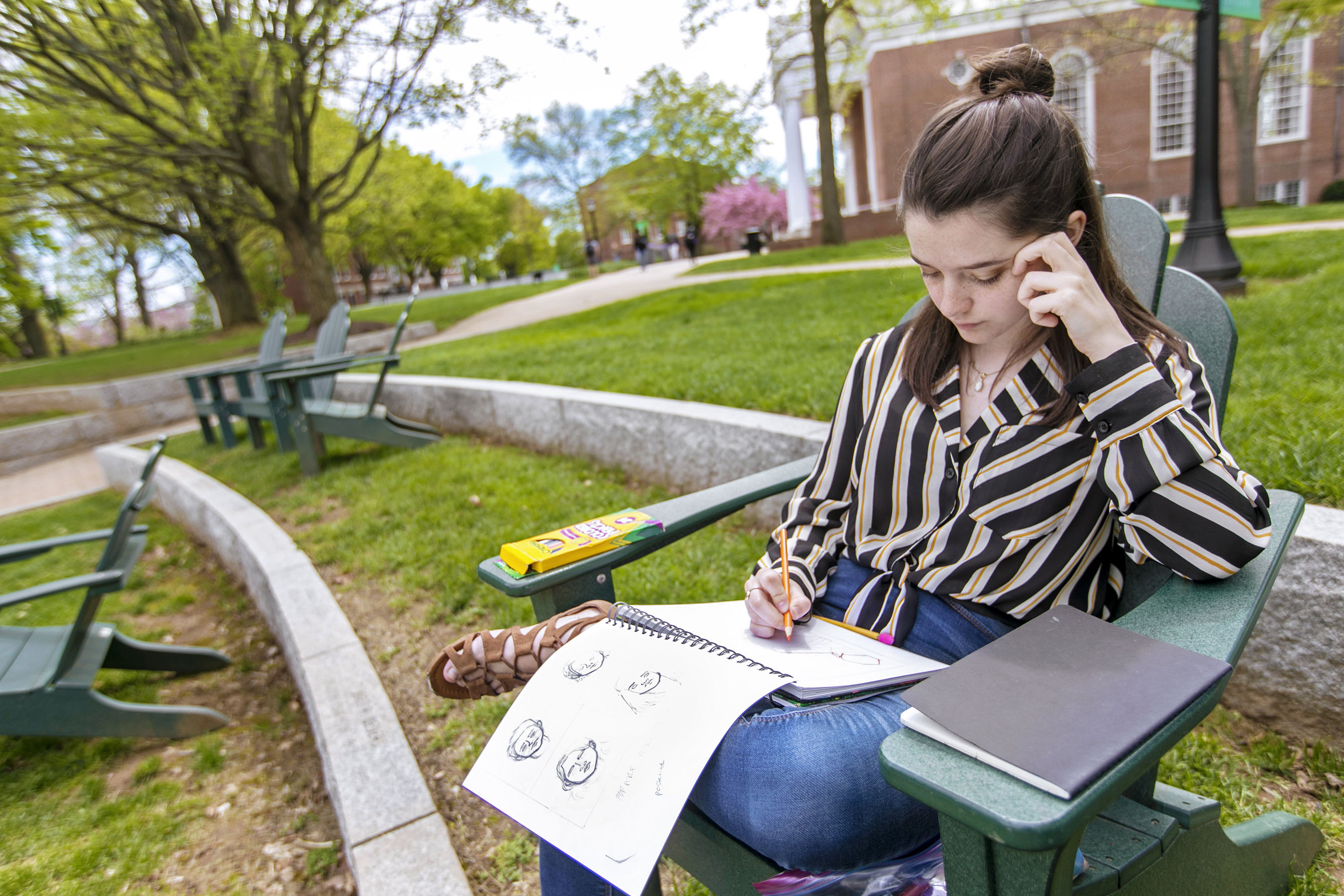 Student drawing in sketchpad while sitting in outdoor chair on campus.