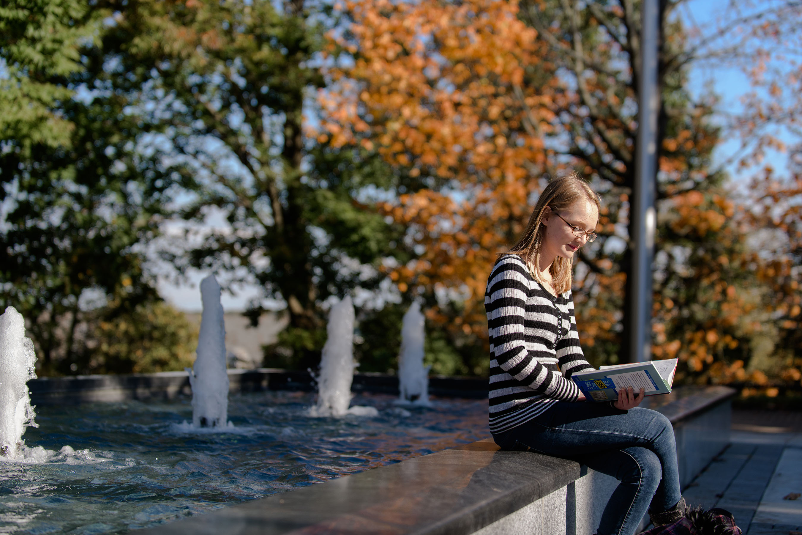 Student reading book sitting on fountain edge on campus.