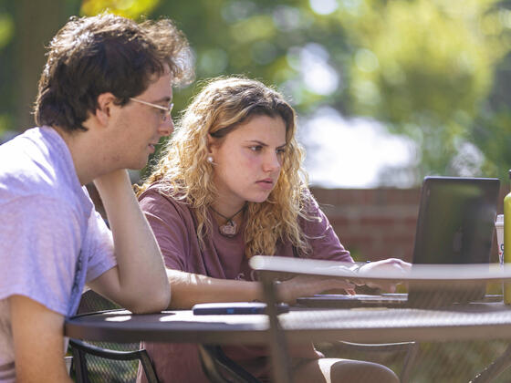 Two students seated together at table outside of Hoover Library looking at computer..