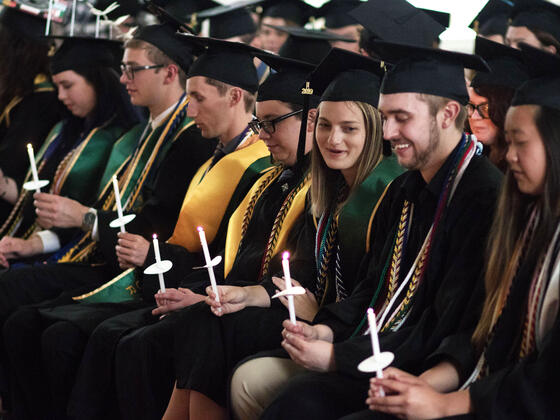 Graduates in cap and gown seated holding candles during ceremony.