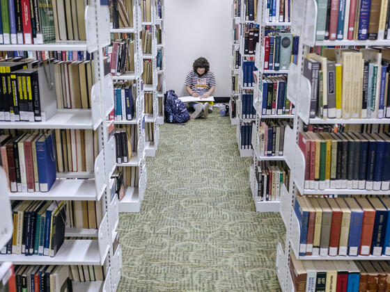 A student sits on the floor in Hoover Library between the aisle of book shelves.