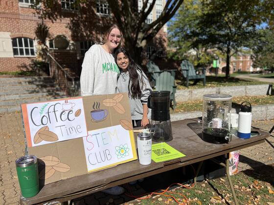 Two students stand at a table with a sign for STEM Club and coffee carafes.