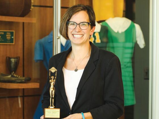 Photo of professor Diane Williams holding a trophy in front of a display of vintage McDaniel sports uniforms.