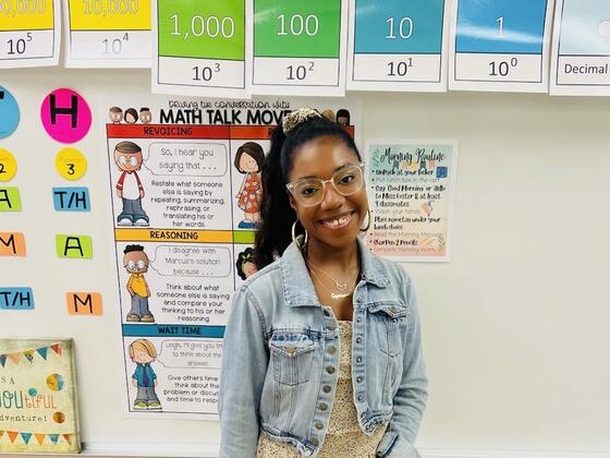 Alumna Amara Foster stands in front of a classroom board with colorful art and lessons on it.
