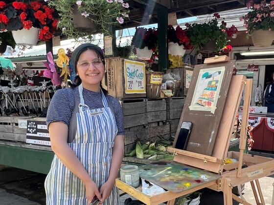 Sarah Mendez at an easel wearing an apron. Behind her is a market stand with flowers.