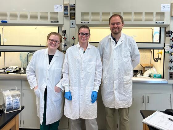 Two students and a professor stand together in the lab, all three wearing white lab coats.