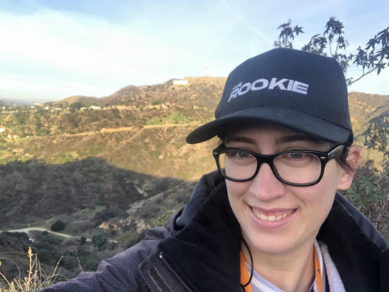 Tricia Meola, a white woman standing in front of the Hollywood sign on a TV set, wearing a hat that says "The Rookie."