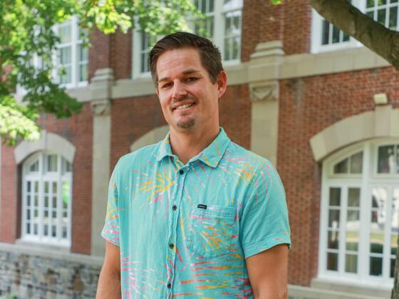 Photo of white male standing in front of a brick building wearing a blue patterned shirt.